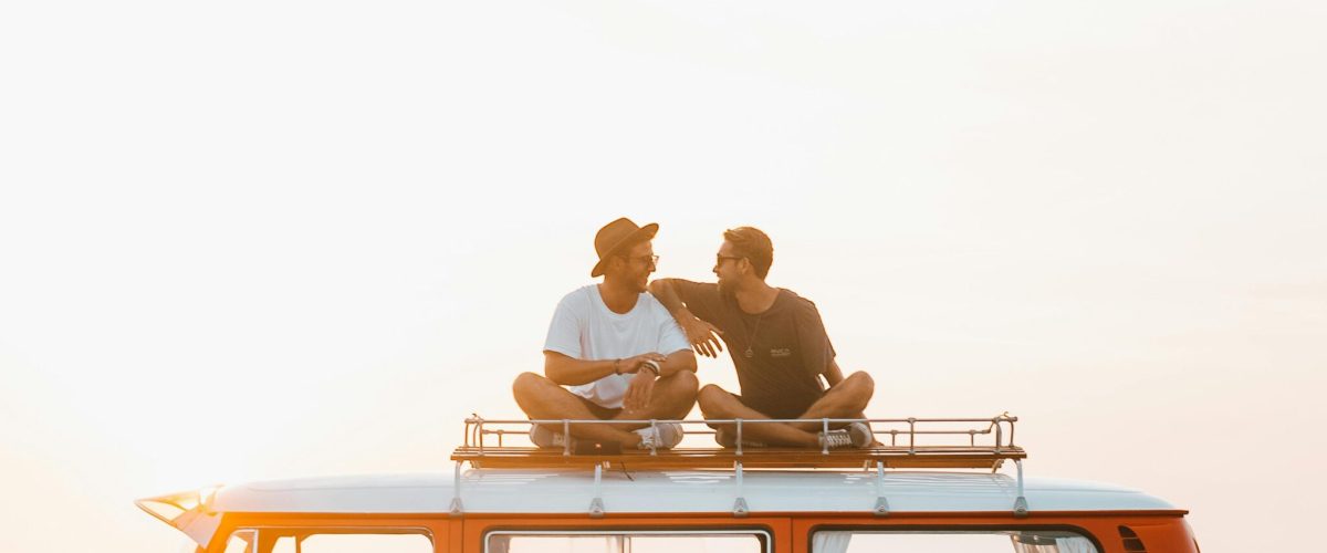 Silhouettes of full body faceless male friends in casual clothes resting on camper van roof and chatting in countryside in sunset time